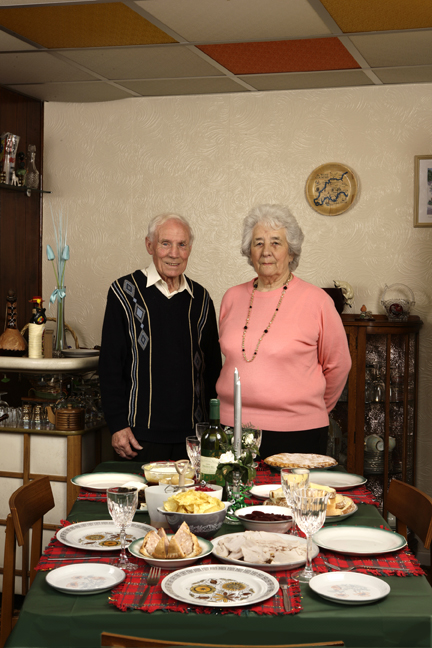 couple John&Darcy stand proudly in front of their table, a documentary image showing an insight into lives, rooms, and special celebratory food,